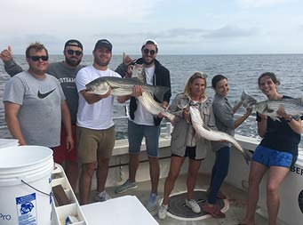 On a cloudy day, seven men and women hold up four striped bass that they caught.