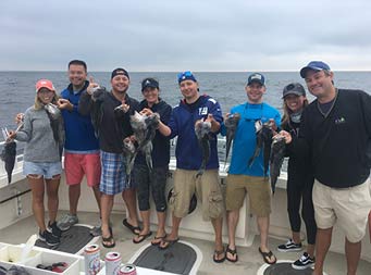With the ocean behind them on a cloudy day, eight men and women stand together and each hold up a sea bass.