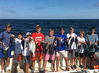 On a sunny day with the dark blue water behind them, 4 young boys ranging in age, stand side by side and each hold up 2 sea bass.