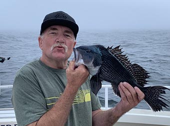 With dense fog behind him, a close up of a man wearing a hat and, smiling, holding a sea bass up to his face.