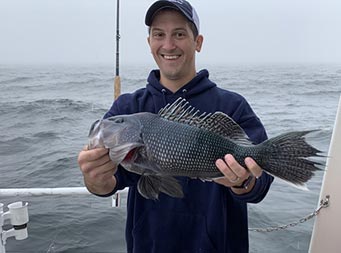 With dense fog behind him, a man wearing a sweatshirt and hat smiles and holds up a sea bass.