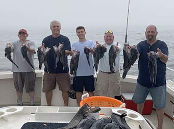 With dense fog behind them, five men smile and each hold up 2 sea bass.
