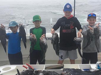 On a cloudy, foggy day, 4 young boys each hold up 2 sea bass.