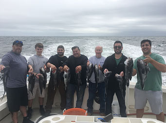 On a cloudy day with the boat's wake behind them, seven men, ranging in age. hold up 2 sea bass each.