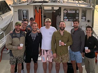 Eight men stand on the boat while still docked at the marina, with the sun setting in the background, and smile for the camera before leaving for their charter.