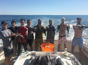 On a clear day with blue sky, all eight men hold up the sea bass they caught.