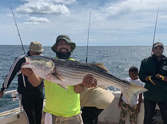 With a man and young boy in the background, a man wearing a bright neon yellow/green shirt in the foreground uses two hands to hold up the striped bass he caught.