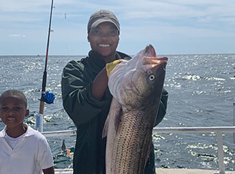 With a young boy beside her, a woman smiles big and holds up striped bass by the gills with two hands.