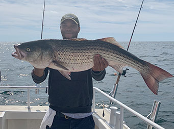 A man wearing a dark color baseball cap and smiling, uses two hands to hold up a striped bass.