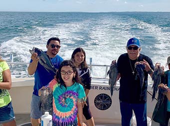With the blue sky and ocean behind them, a group of 7 ranging in age, smile and hold up sea bass and porgy (scup).