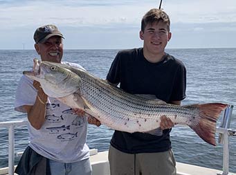 An older man smiles next to a young man also smiling, and holding up the striped bass he caught.