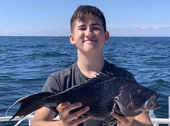 With the blue ocean behind them, the camera takes a close-up of a proud young man holding up the sea bass he caught.