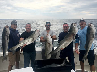 On an overcast day, 6 men smile for the camera and each hold up a striped bass.