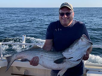 A man holds up a striped bass with both hands and smiles.