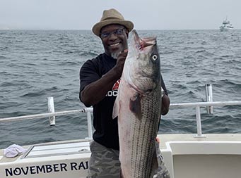 On an overcast day, a man holds up a striped bass with both hands and smiles.