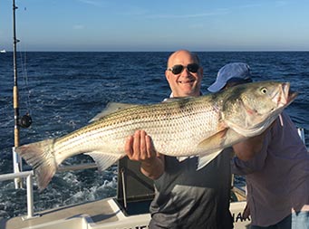 A man holds up a large striped bass with both hands and smiles.
