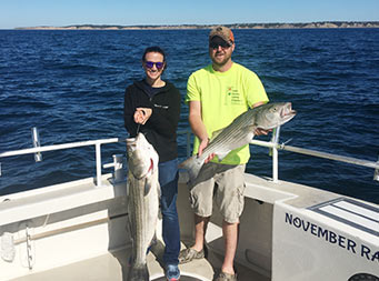 With a clear blue sky and the dark blue ocean behind them, a man wearing a bright yellow neon long t-shirt, and a woman wearing a black long sleeve shirt each hold up striped bass.
