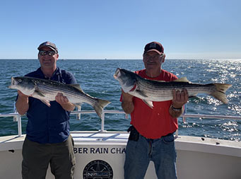 With a sunny clear blue sky above them, two men each hold their own striped bass and smile.