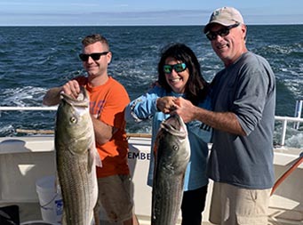 A man wearing an orange shirt and sunglasses holds up a striped bass with 2 hands, and a woman and another man hold up their own striped bass together.