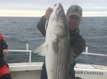 On an overcast day, a man uses both hands to hold up the striped bass he caught.