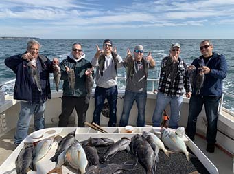 On a sunny sky with some clouds, six men each hold up 2 sea bass and smile.
