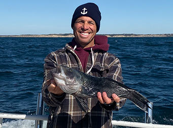 A man wearing a hat and flannel smiles and holds up a sea bass with 2 hands.