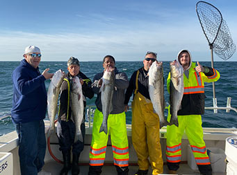 On a sunny day with light clouds, 5 men wearing winter attired each hold up a striped bass.