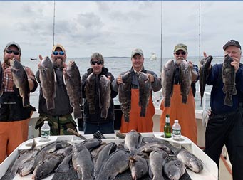 On an over cast, cloudy day, 6 men wearing fall attire, and some wearing orange fishing waders, each hold up 2 sea bass with more laid out in front of them on the filet table.