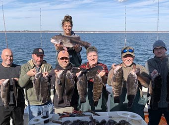 On a 
 sunny day with few clouds, Captain Jill stands in the background on the edge of the boat holding up one blackfish, while 6 men in front of her each hold up 2 sea bass, and more sea bass laid out on the filet table.