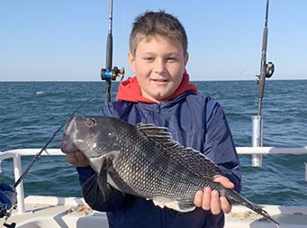 With the sun lighting a clear sky, a tween-age boy holds up a sea bass for the camera with both hands.