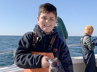 On a clear, sunny day, a a tween-age boy wearing orange Grundens fishing waders smiles and holds up a sea bass.