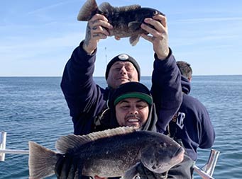 A man in the foreground smiles for the camera and holds up a large sea bass, while his friend stands directly behind him and holds up a much smaller sea bass over his head.