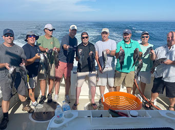 With a partly cloudy sky in the back, 9 men standing together each hold up 2 sea bass for the camera
